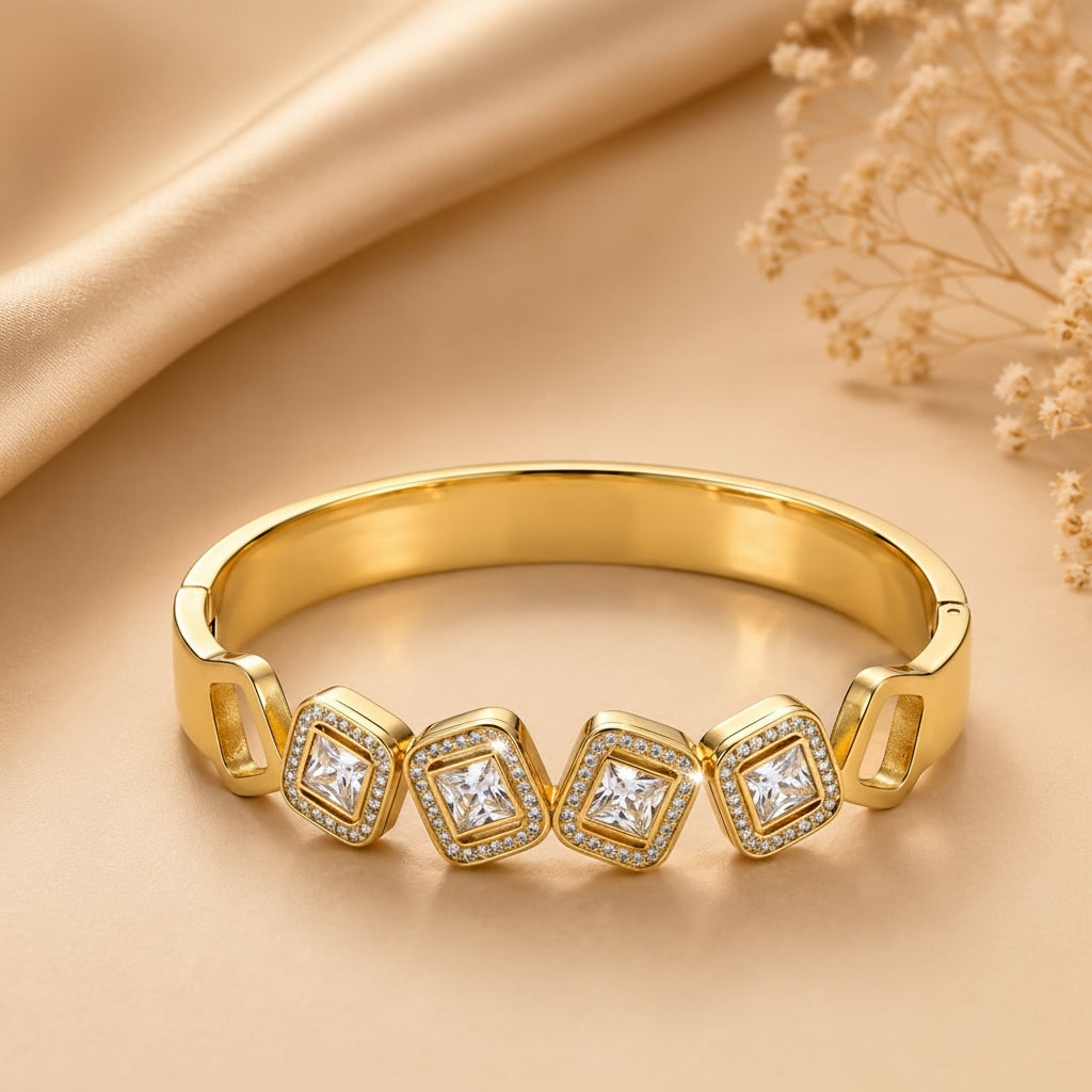 A polished yellow gold band ring featuring four square-cut clear gemstones in bezel settings, accented with small round stones, displayed on a beige satin fabric background with dried flowers.